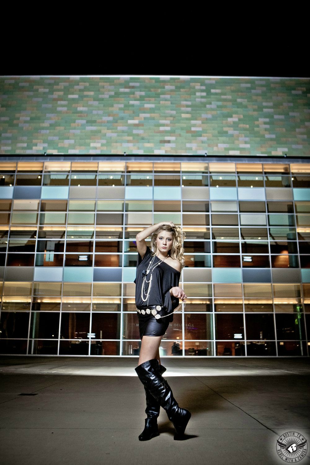 hot blond female model with curls and short black vinyl skirt, black knee high boots and off the shoulder black blouse with medallion chain belt and long necklaces in front of the Long Center with a glass wall with green, light green, gold and brown tiles in background